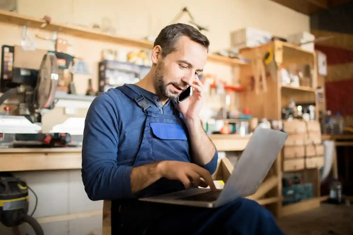 Handwerker telefoniert in seiner Werkstatt mit einem Kunden, vor sich den Laptop